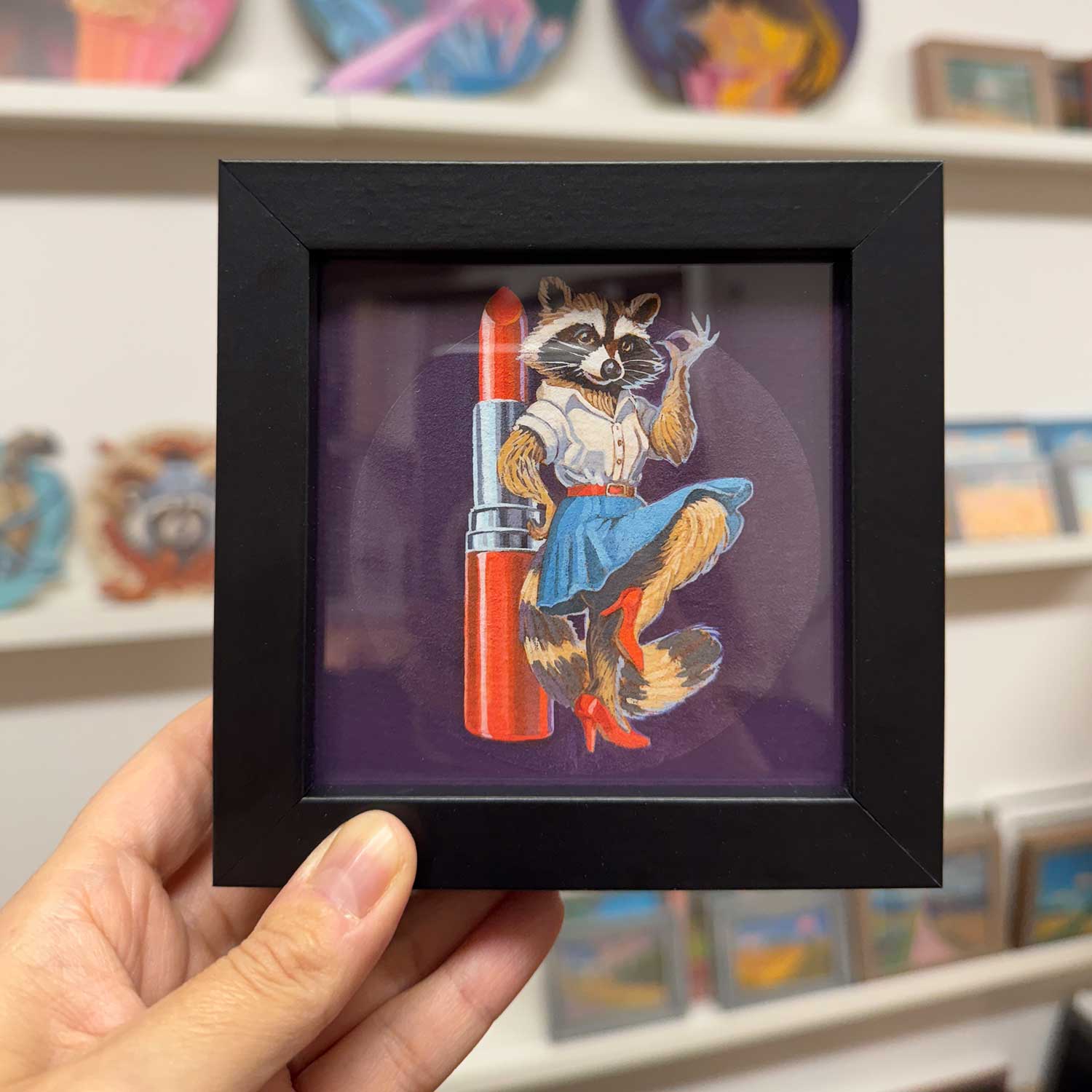 Cute raccoon in a dress holding lipstick in a trendy black frame, perfect for adding fun to any space!