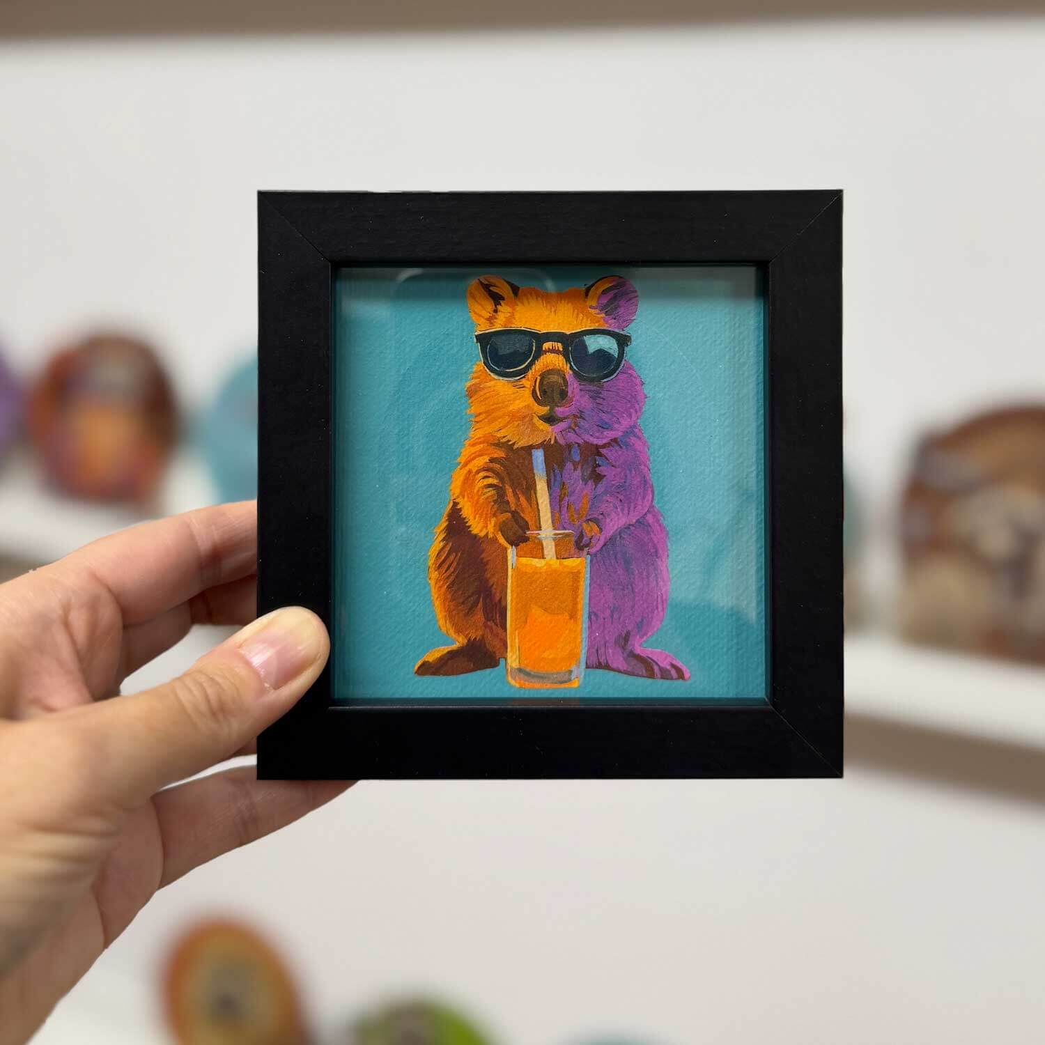 Quokka art print in a black frame, holding a drink, bringing fun to any space! Perfect for home decor.