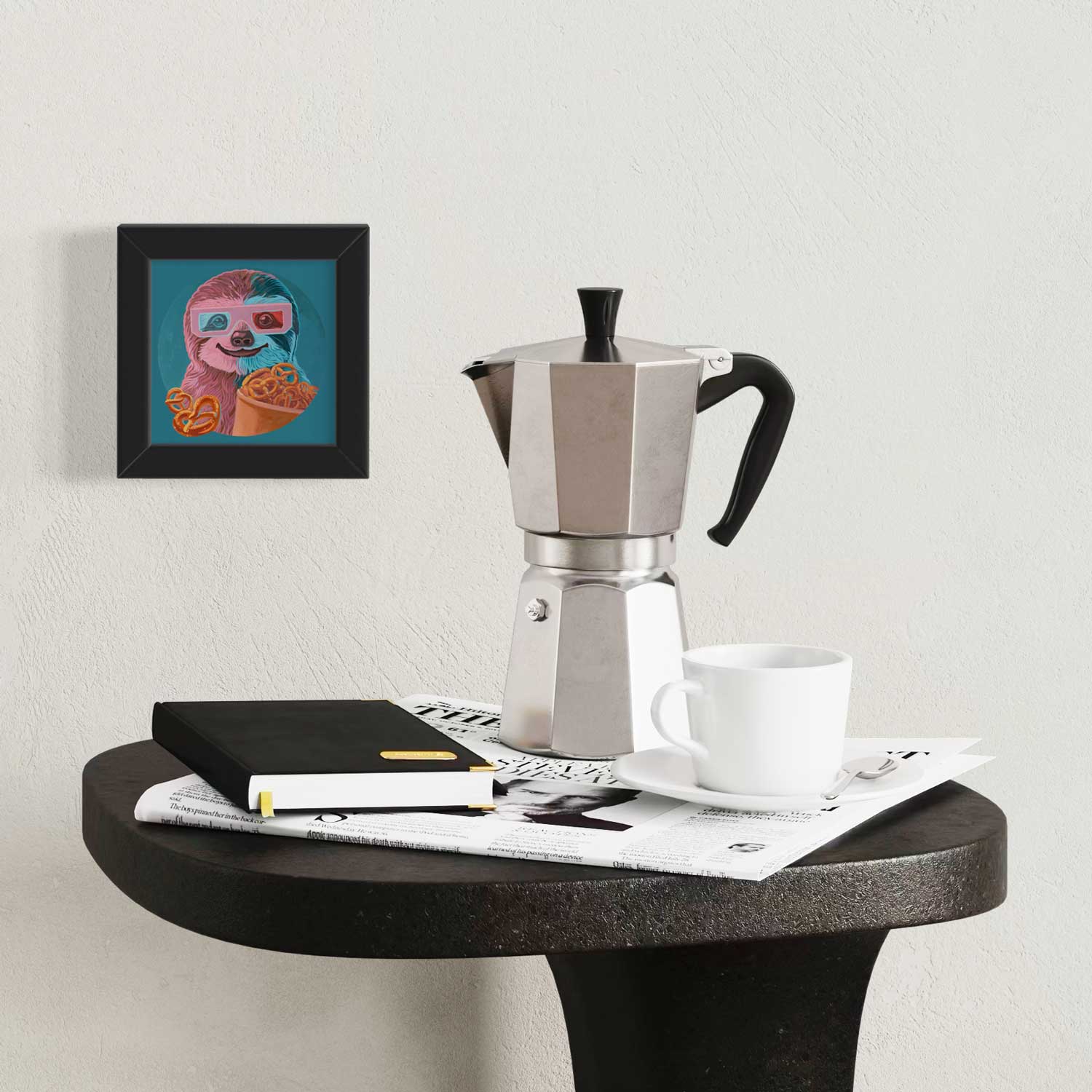 Framed sloth art print beside coffee maker and notebook on a stylish table, brightening up the cozy space.