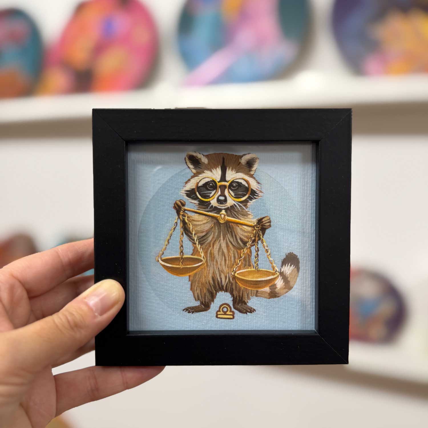 Cute raccoon holding scales, framed 10x10 cm art print for Libra zodiac, perfect for decorating small spaces.