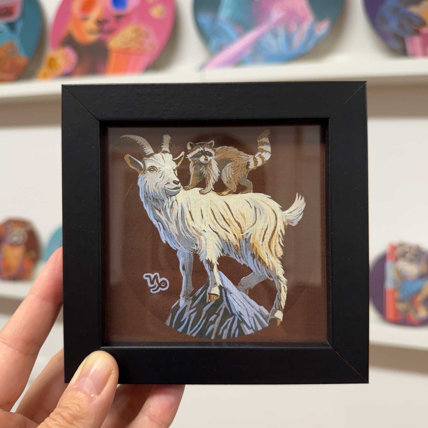 Pocket Art print featuring a raccoon on a goat, framed in a stylish black frame, adding charm to any space.