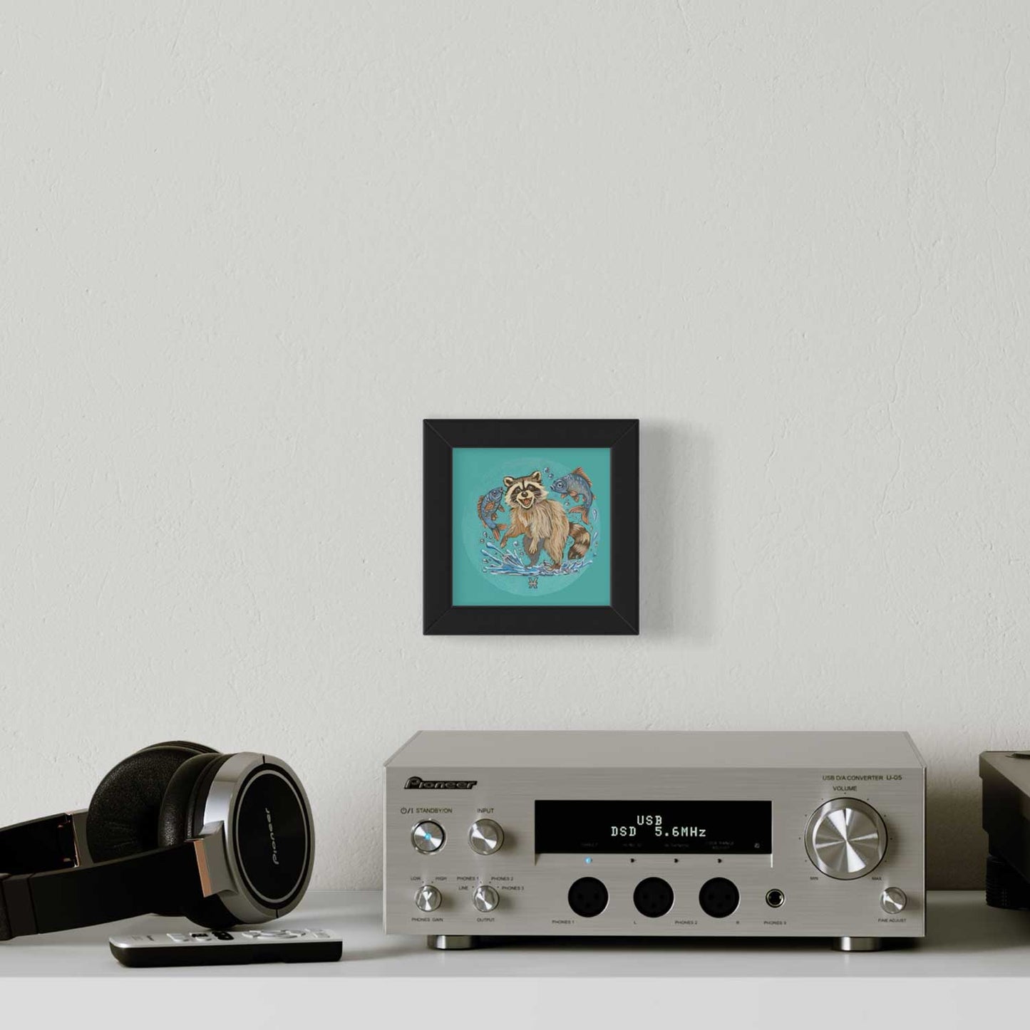 Cute raccoon fine art print framed in black, brightening up a modern desk setup with headphones and a stereo.