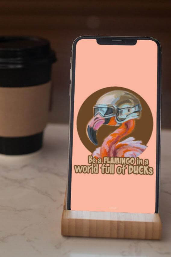 Handy mit Flamingo-Wallpaper und Spruch "Be a flamingo in a world full of ducks", Holzständer, Kaffeebecher im Hintergrund.